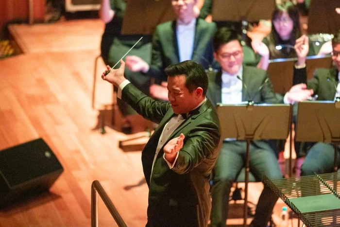 Adrian Chiang conducting on stage, baton raised, with the orchestra behind him